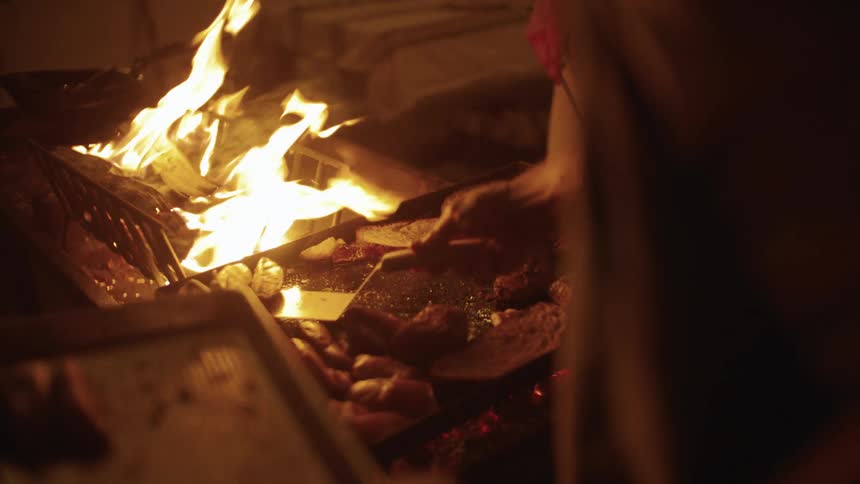 A person grills meat over an open flame at night