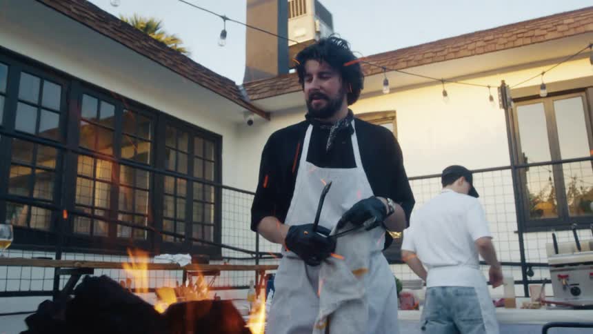 A chef stands by a grill with flames rising preparing food outdoors