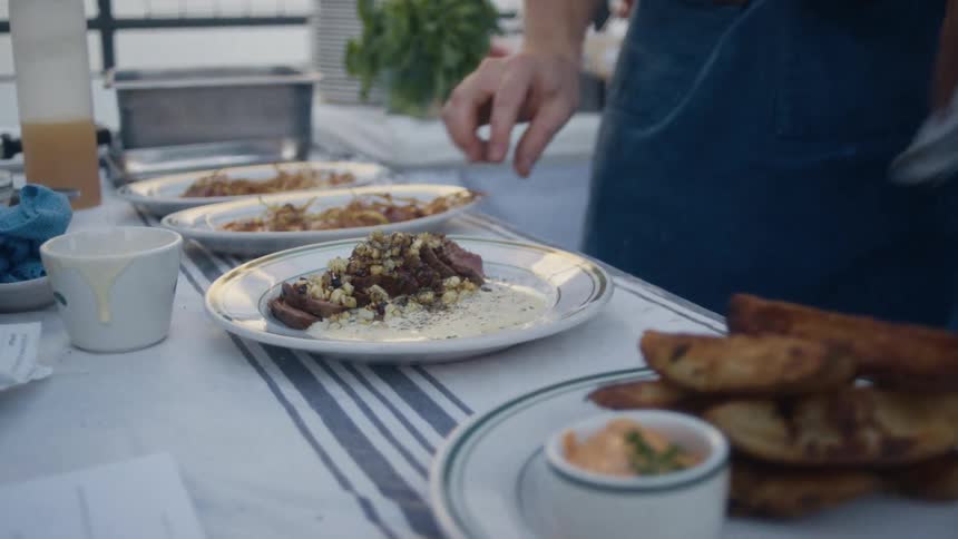 A person in an apron is plating and presenting gourmet food on a table outdoors