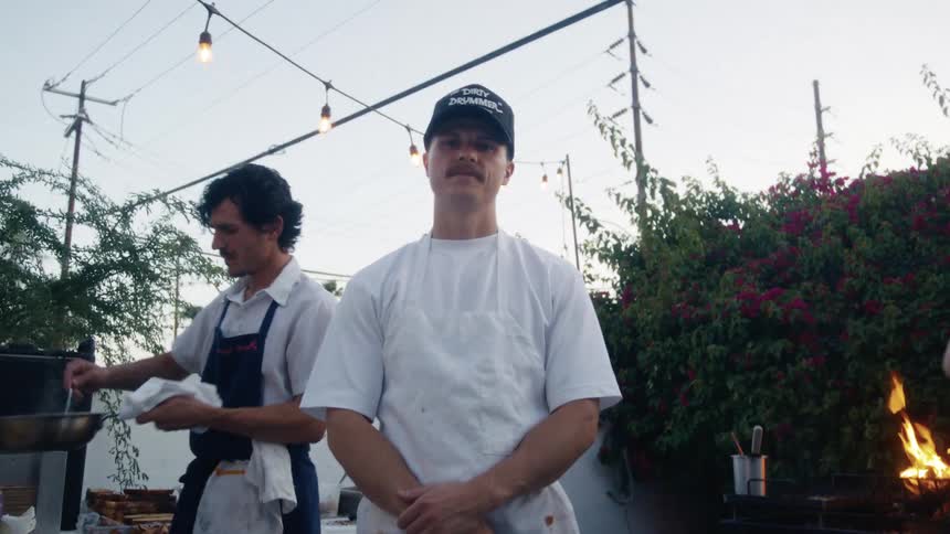 Male chef stands outdoors while others cook near him on a summer day