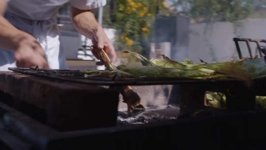 A person grills corn and leafy vegetables over an open flame on a backyard barbecue