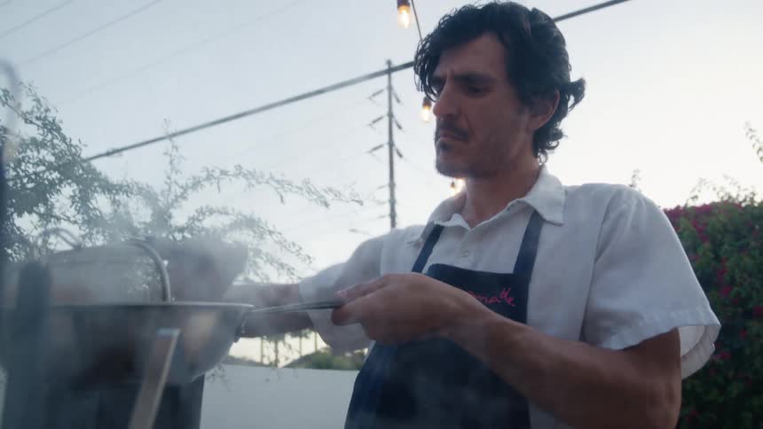 A chef strains steaming pasta in a smoky open air kitchen setting