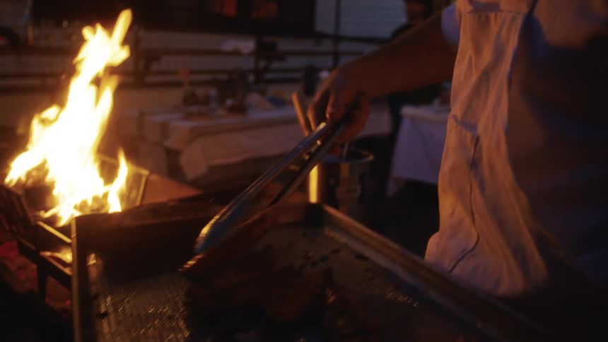 A chef cooks food on a tray near an open flame in a dimly lit outdoor setting at night