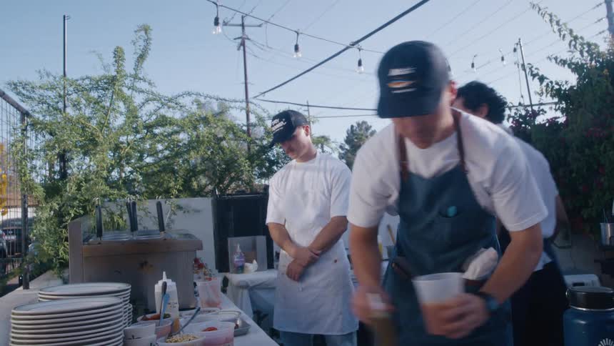 Several chefs working outdoors in what appears to be a casual food preparation area they are focused on mixing and preparing ingredients in containers suggesting a catering event or an outdoor kitchen