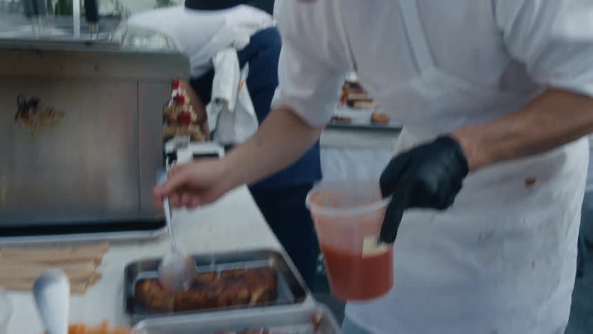 A person wearing black gloves and a white shirt meticulously spreading a red sauce onto two pieces of toasted bread on metal trays other food items and containers are visible on the table indicating food preparation for an event