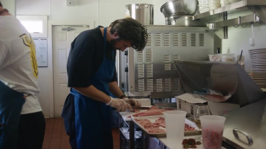 A chef slices meat on a cutting board in the kitchen with another man standing behind him