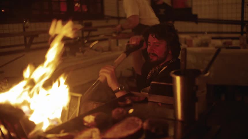 A chef works near an open flame grill in a dimly lit kitchen setting