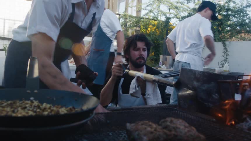 A man stirs the fire with a shovel as other men cook
