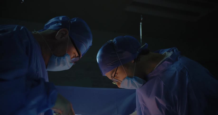Doctors in a theater room wearing blue coats headbands and nose masks perform surgery on a patient