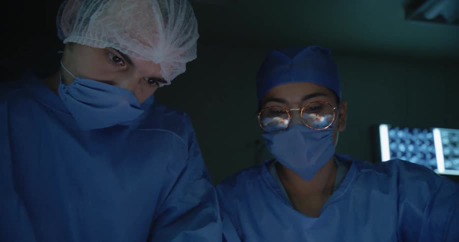 Two surgeons in blue scrubs and masks focus intently during an operation under dim surgical lighting