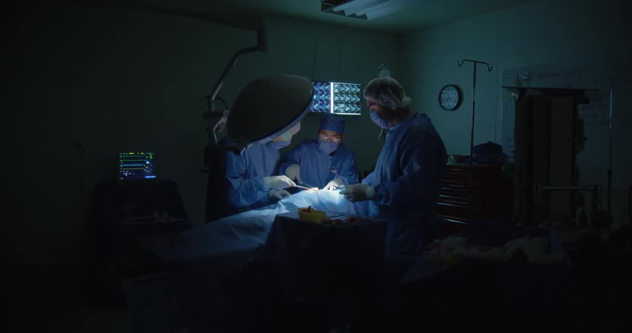 Three surgeons perform an operation under focused lighting in a dark operating room