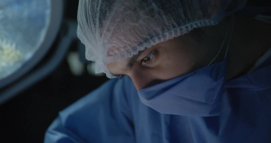 A close view shows a person in a surgical cap and mask looking down with focused eyes against a dark background
