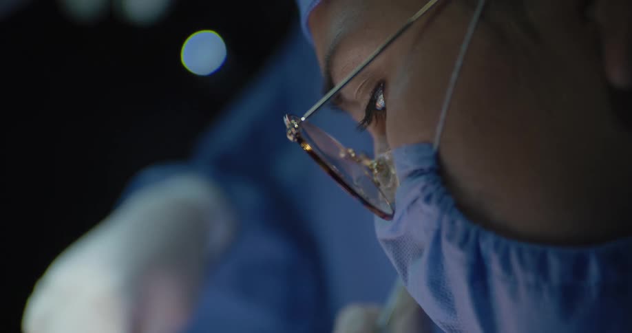 A person wearing blue scrubs and a face mask looks down with intense focus with a bright reflection from surgical loupes visible on their glasses