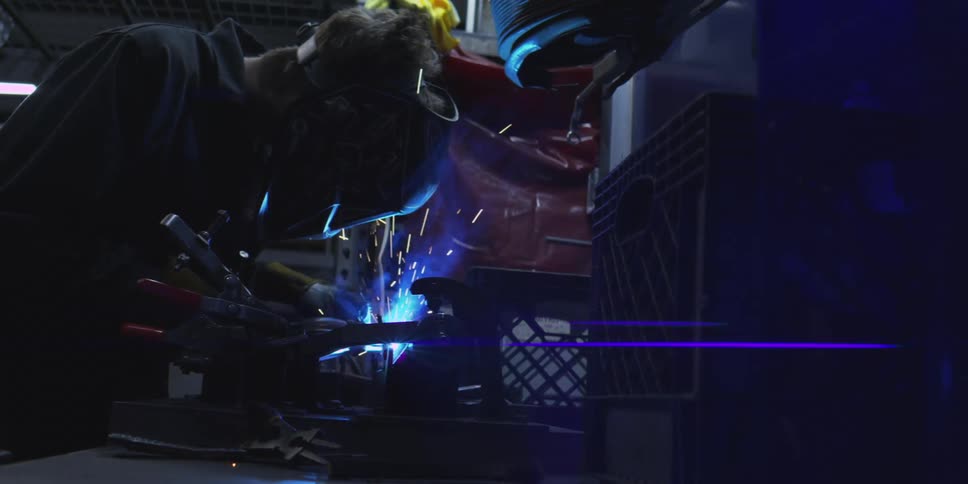 A man wearing welding face gear welds a piece of metal inside a workshop