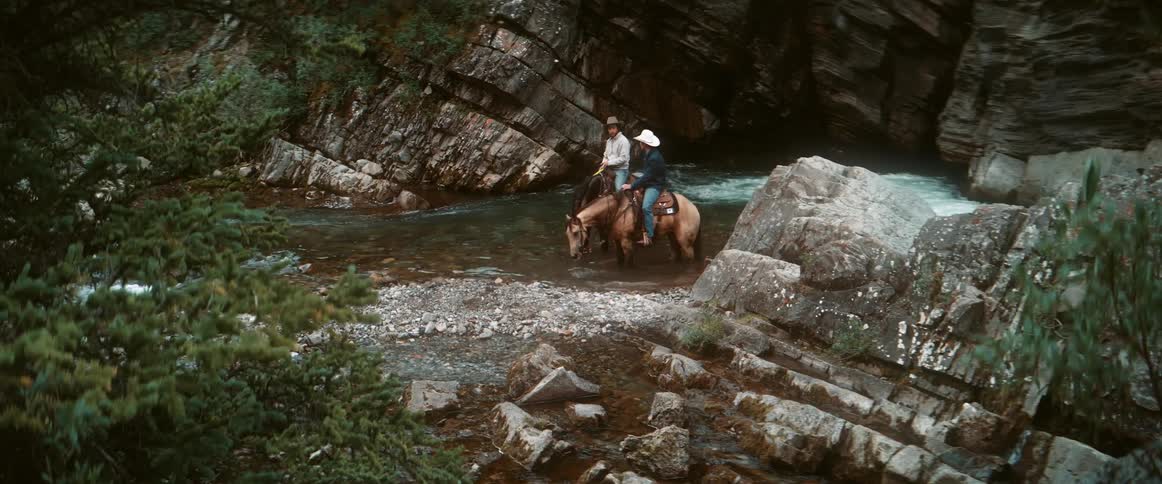 Two cowboys riding horses stop in the stream to let the horses drink
