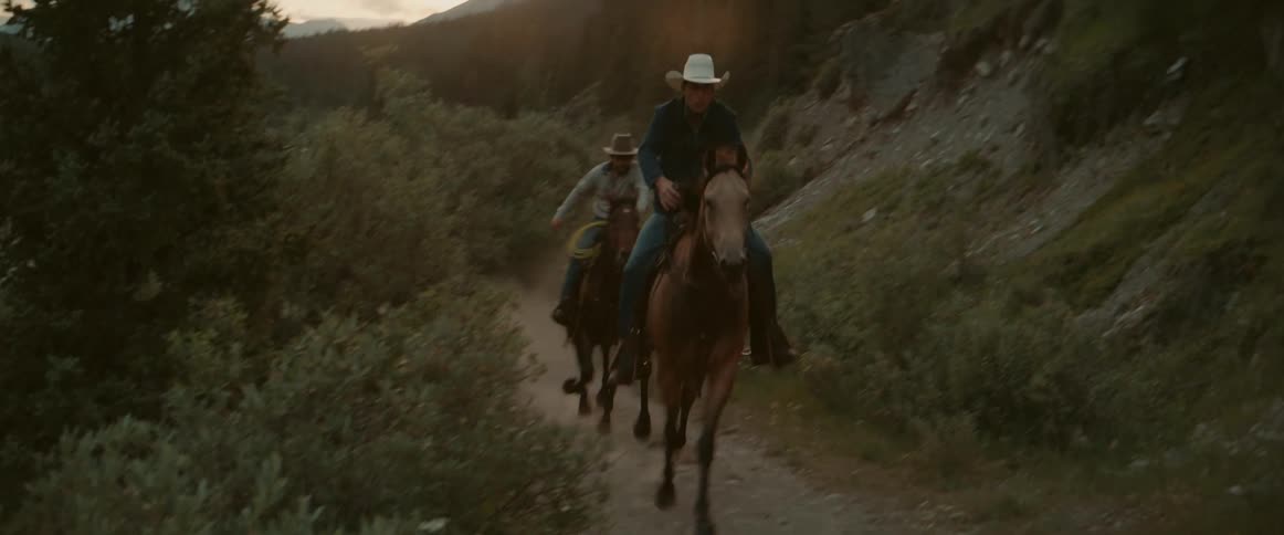Two men wearing sun hats ride horses quickly into the woods at midday