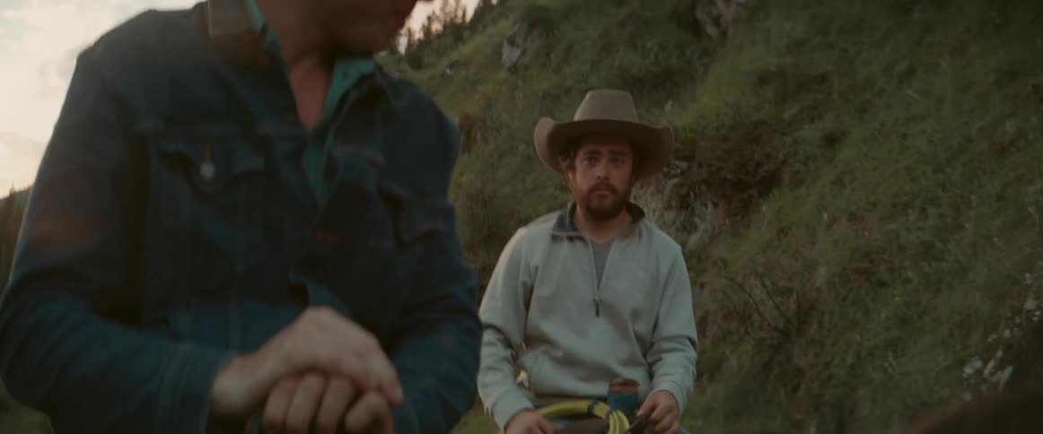 Two men on horseback with one in the foreground wearing a denim jacket and a white cowboy hat that he momentarily takes off smiling the other man in a grey top and darker cowboy hat looks on as they ride horses through a landscape with hills