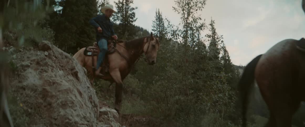 Two men ride horses down a rocky hill surrounded by trees at midday