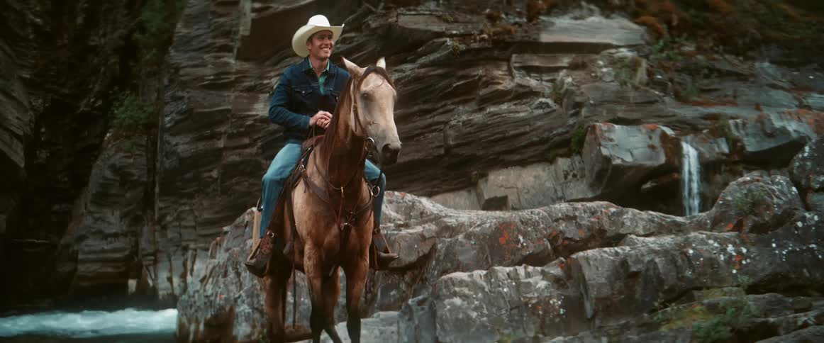 A cowboy wearing a white hat and denim outfit rides a tan horse along rocky terrain near a small waterfall and rugged cliffs
