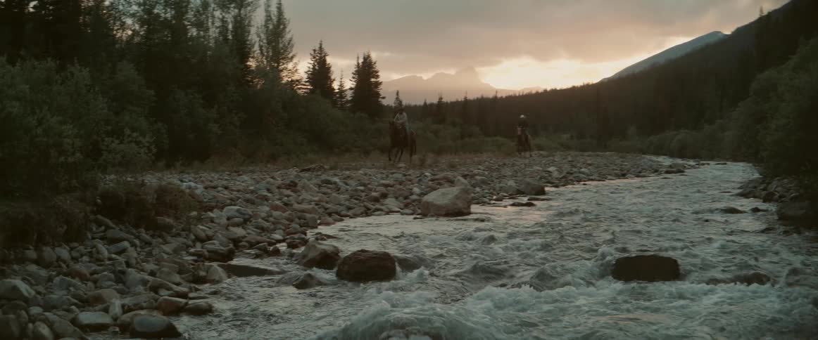Two horseback riders travel along a rocky riverside at sunset with a fastflowing stream in the foreground and forested mountains silhouetted in the distance