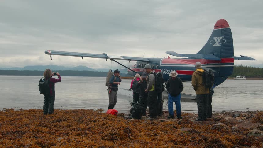 Several people stand on a rocky shoreline near a floatplane with seaweedcovered rocks underfoot and mountains in the distance