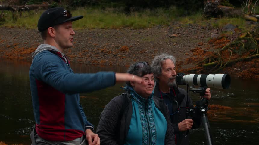 Man points into the distance as couple watches and camera with long lens is mounted on tripod