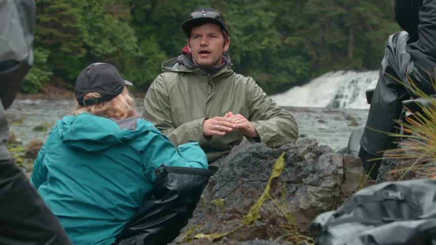 A group of people dressed in outdoor gear sit and talk near a rocky riverside with a waterfall and lush forest in the background