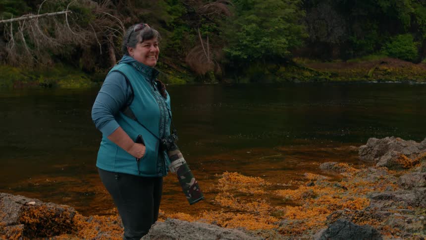 A smiling woman stands on a rocky shoreline beside a calm river dressed warmly with a camera and long telephoto lens hanging from her neck