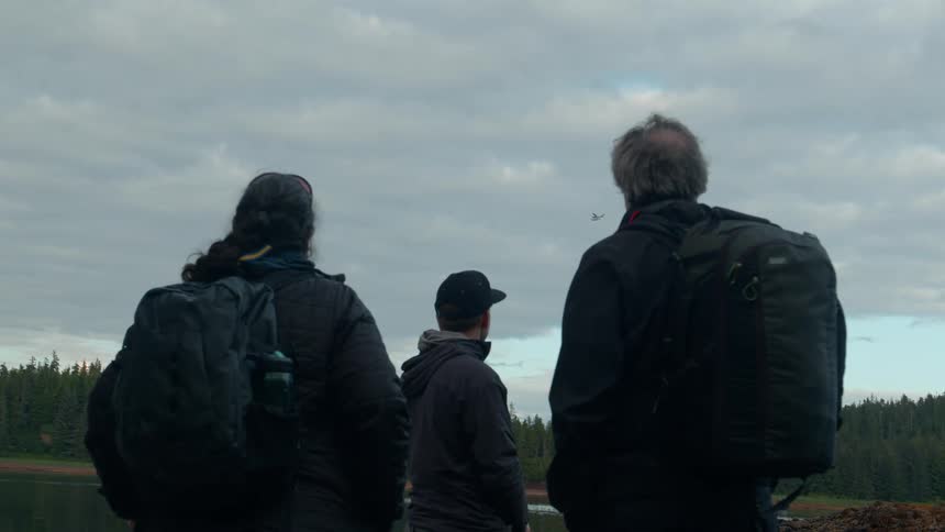 Three individuals all wearing backpacks and facing away from the camera looking out over a body of water towards a forested landscape under a cloudy sky a small plane can be seen flying in the distance between them
