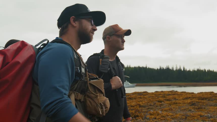 A group of hikers dressed in outdoor gear and carrying large backpacks walk along a rocky seaweedcovered shoreline under a cloudy sky near calm coastal waters