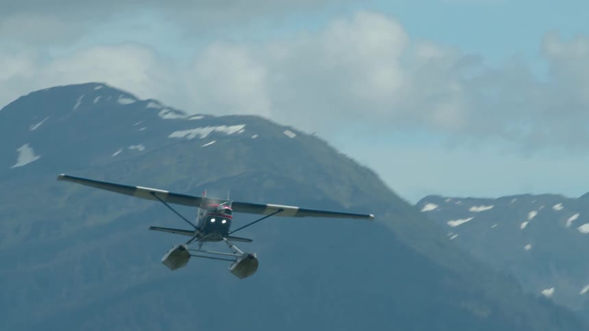 A seaplane flies low just above a woodland heading toward a lake with a mountain view in the background at midday