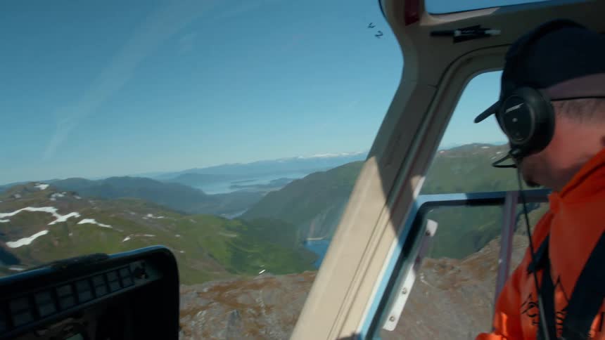 Man in cap and headphones inside a helicopter flying over mountains