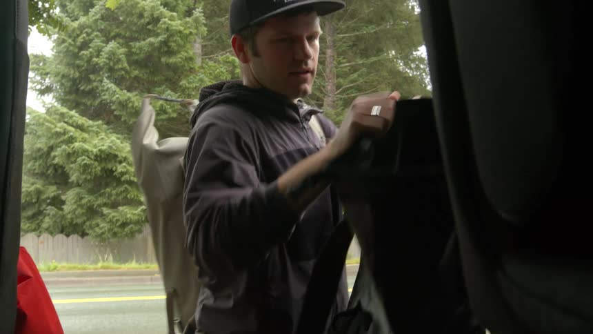 A man who appears to be packing or organizing gear inside what looks like the back of a vehicle possibly an suv or van given the anglehes wearing a dark hoodie jeans and a baseball cap he has a large lightcolored possibly waterproof dry bag
