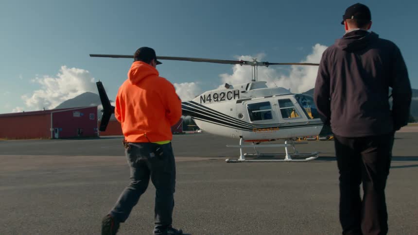 Two adult men among them one is wearing a orange hoodie and a cap one is wearing a black hoodieand a cap are walking towards a helicopter