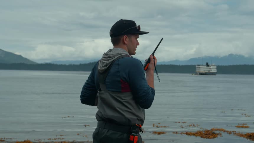 A man in fishing waders stands on a rocky shore and talks on a walkie talkie as a seaplane lands on the water behind him