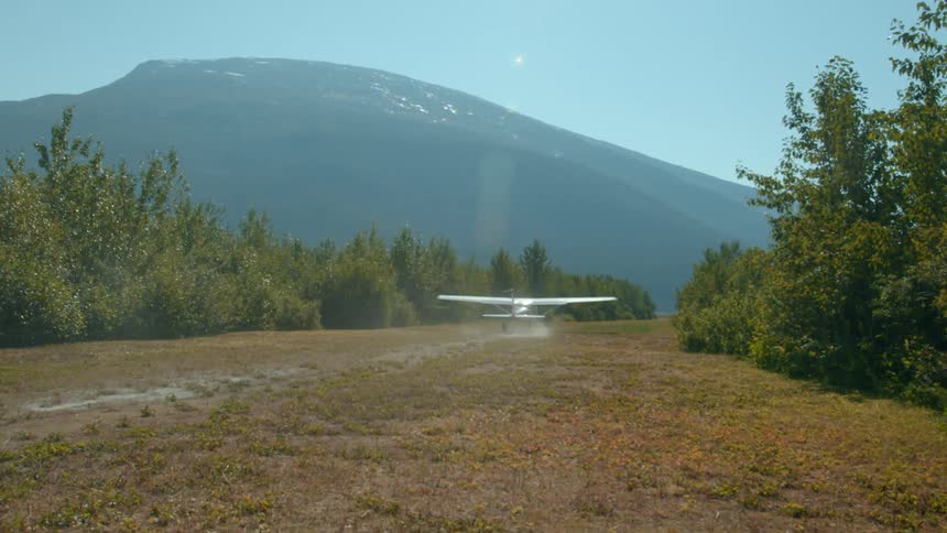 A small plane takes off from a grassy runway surrounded by trees with a towering mountain rising in the background under a clear blue sky
