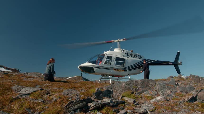 A helicopter prepares for takeoff on rocky terrain as two people stand by one looking nervous