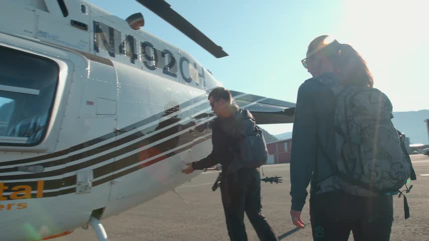 A man and a young woman both carrying backpacks place them into the internal compartments of a helicopter at the airport