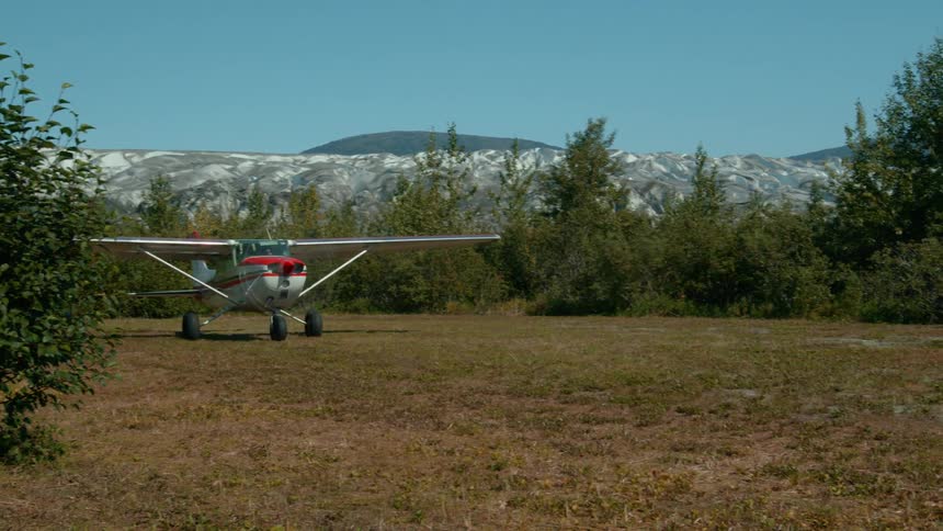 A small propeller plane rests on a rustic grassy airstrip set against the dramatic backdrop of a massive glacier and forested mountains