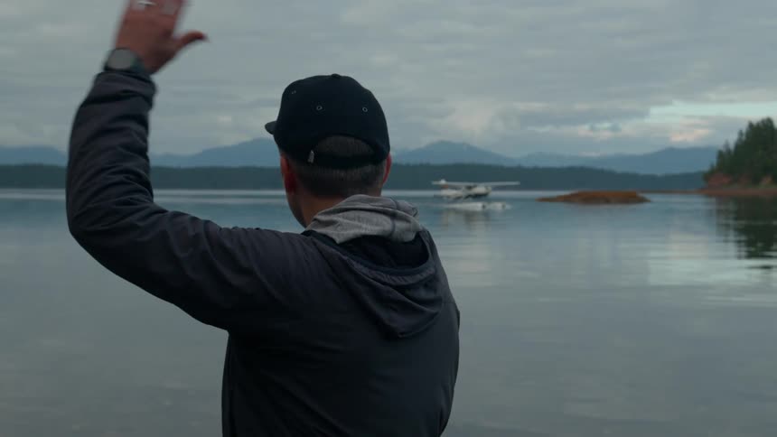 Man standing on the shore waves as a seaplane moves through the water near a small islet on an overcast day