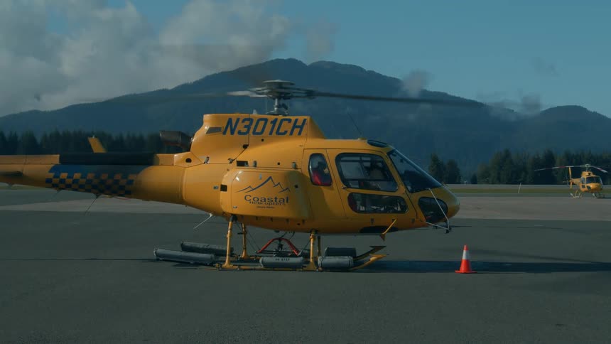 A yellow helicopter sits parked on an airstrip with mountains in the background