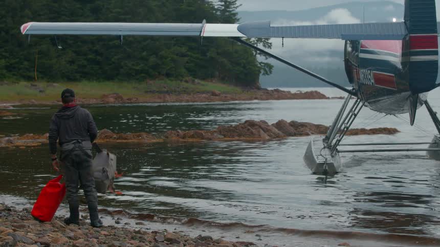 A person wearing a backpack and holding a red bag stands on a rocky shore looking towards a floatplane docked in the water the scene suggests an adventure or travel in a remote natural setting
