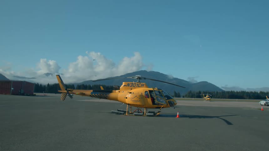 A bright yellow helicopter marked n301ch sits on a tarmac with mountains and clouds in the background alongside another helicopter and a parked vehicle