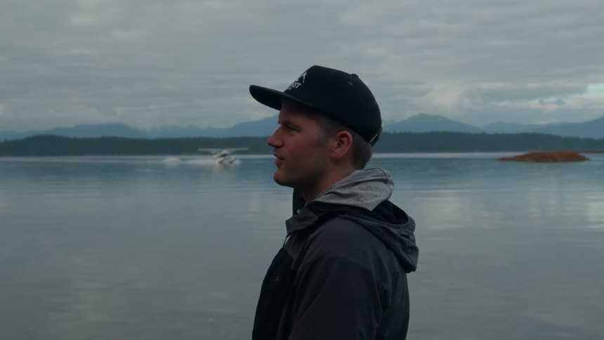 Man in jacket and cap on the shore of a lake with mountains on the horizon