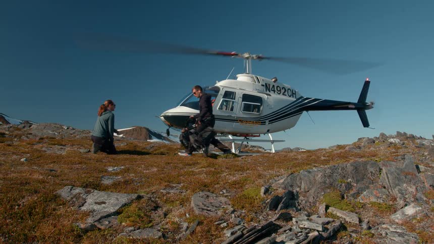 Man and woman on a mountain with a helicopter next to them with the blades on