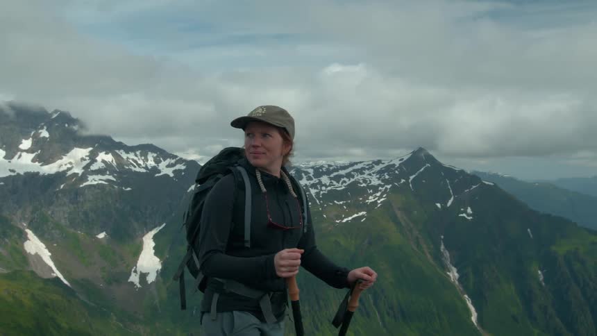 A young teenage girl stands on top of a mountain during a hike at midday wearing a sweater and hat carrying a backpack and holding hiking sticks with surrounding mountain peaks covered in snow