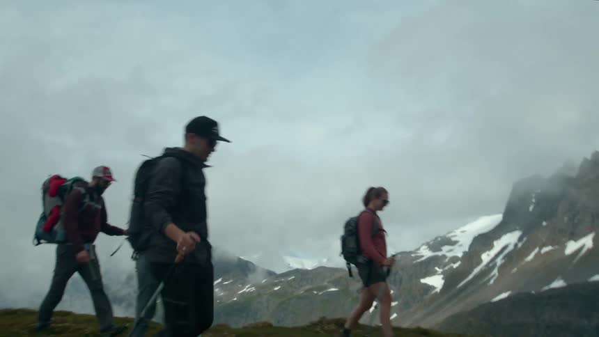 Backpackers hike on a grass covered field across a valley from a snow dappled mountain on a cloudy day