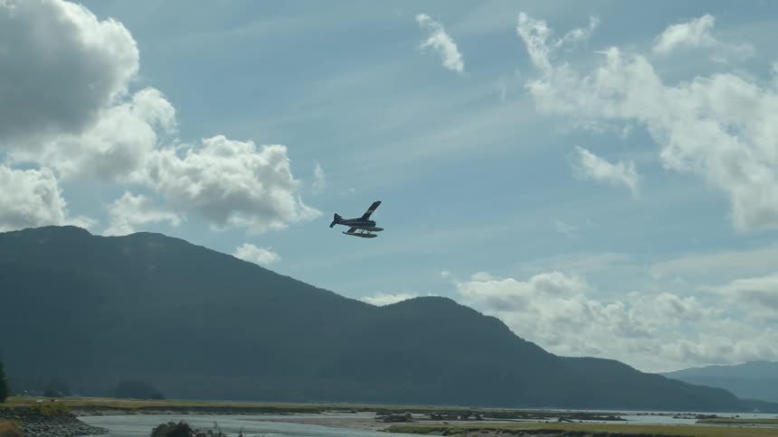 A seaplane takes off flying low over a wide river valley with forested mountains in the background under a partly cloudy sky
