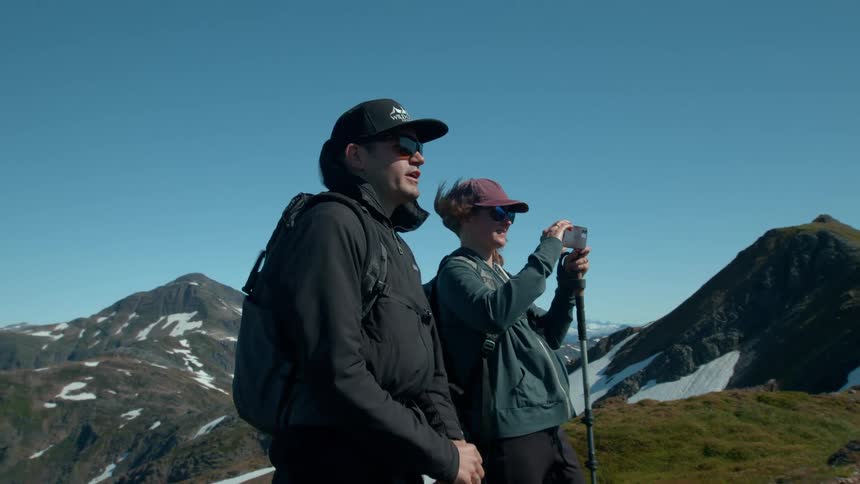 A couple wearing caps and carrying backpacks stand on a mountain ridge filming with a phone with a vast expanse of snowy peaks and valleys behind them under a clear blue sky