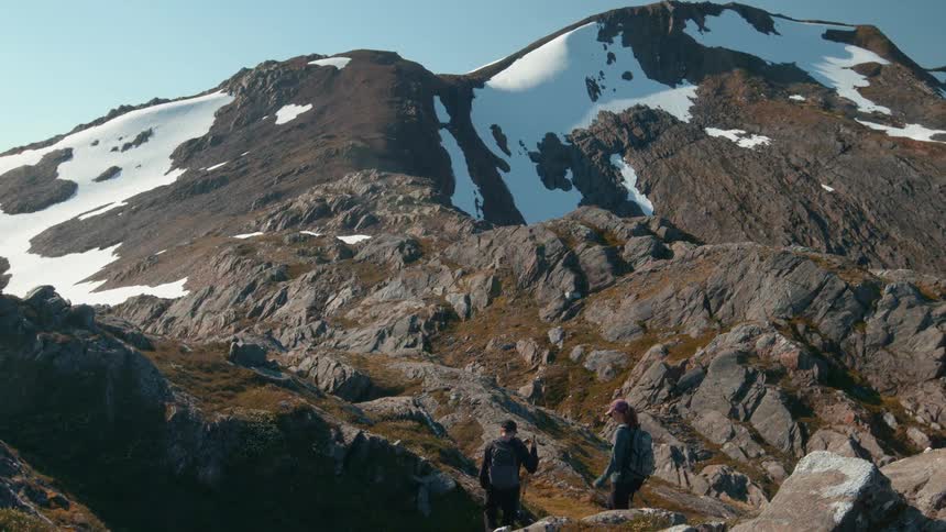 Hikers walk across rocky terrain with snow covered mountains behind
